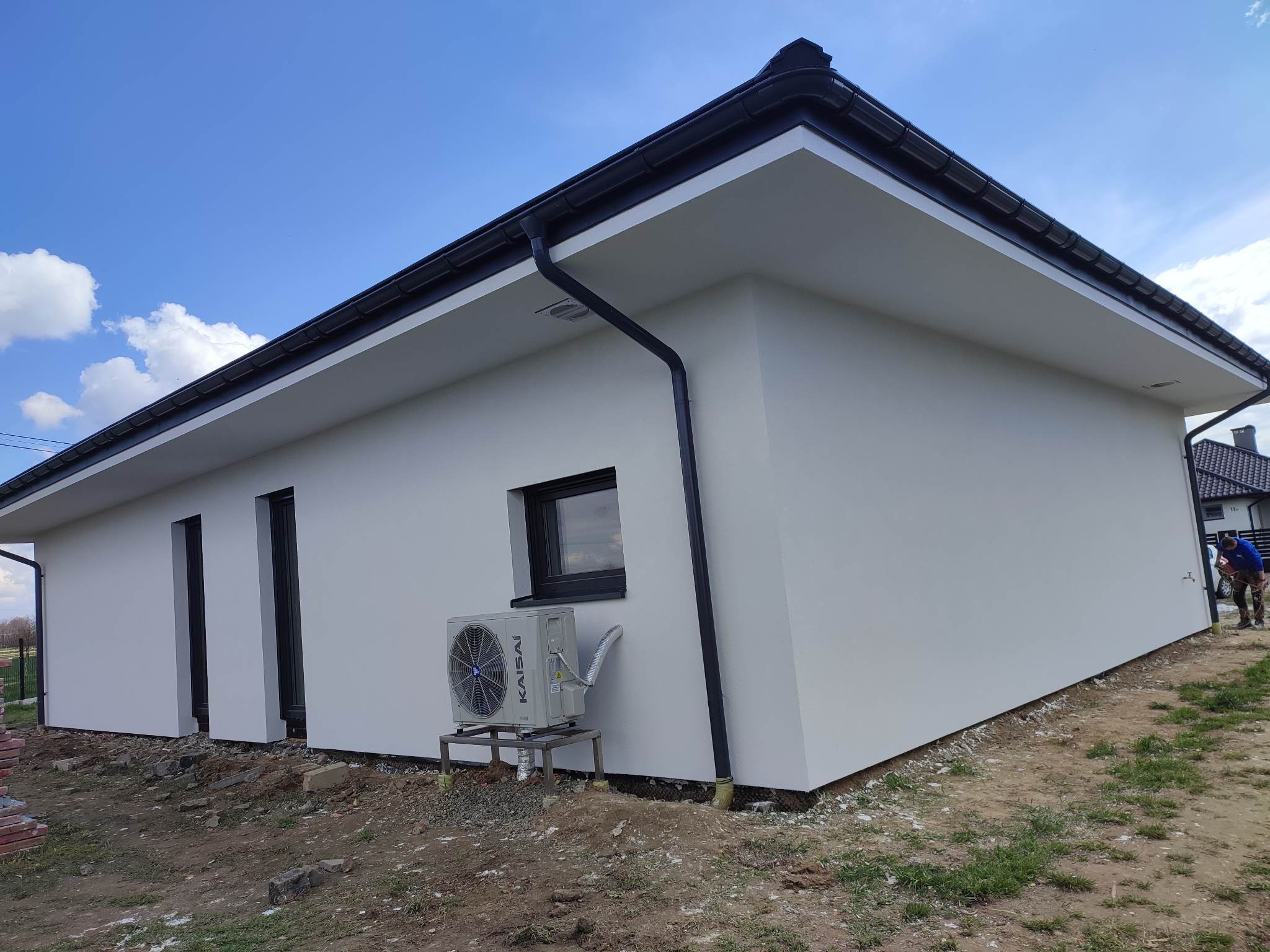 Świeżo otynkowany, biały budynek z czarną stolarką okienną i orynnowaniem, klimatyzator Kaisai na zewnątrz, widoczny fragment niezagospodarowanego terenu.