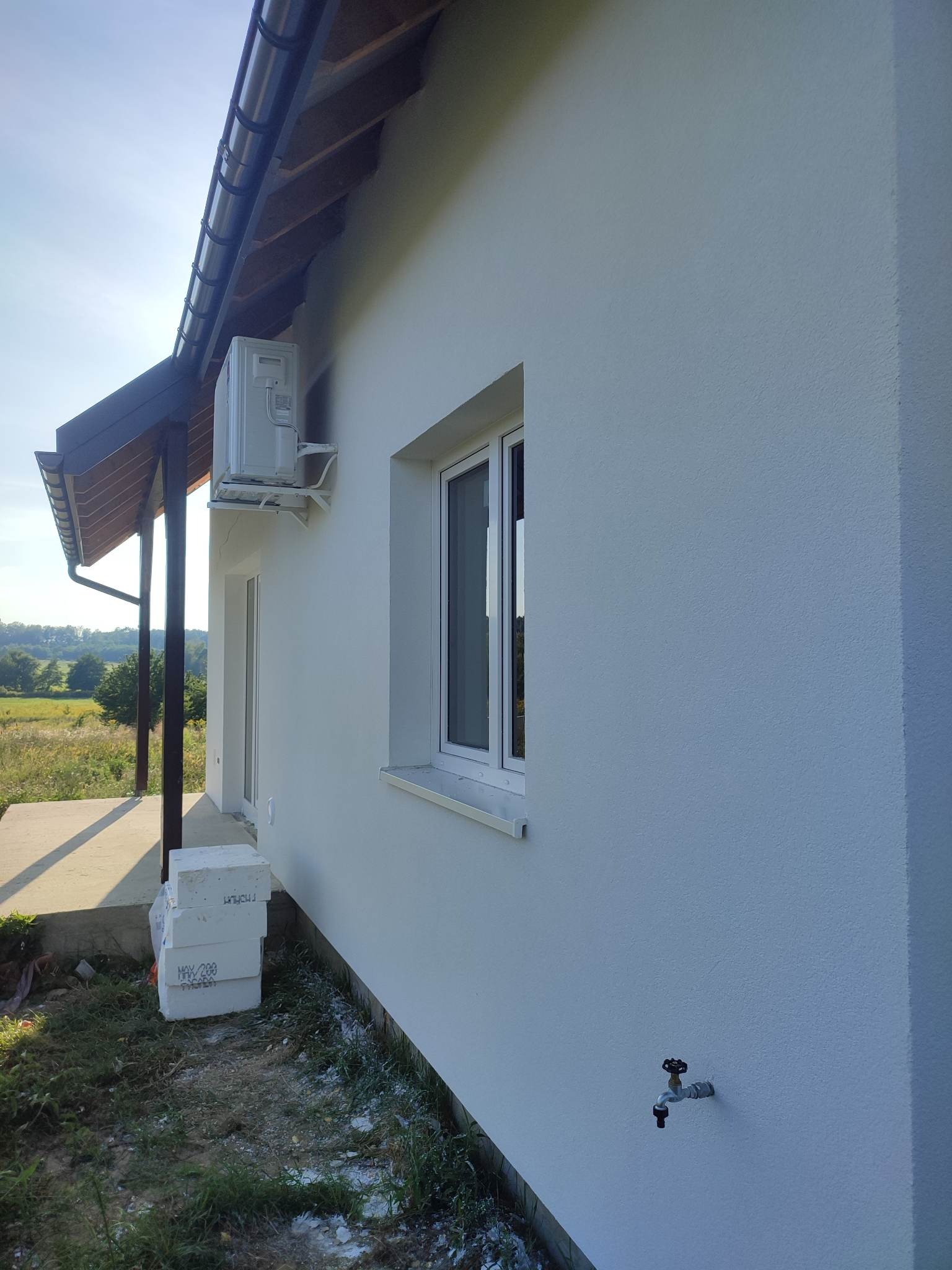 Świeżo otynkowana, biała elewacja domu jednorodzinnego z widocznym oknem, klimatyzatorem i stosem płyt styropianowych przy fundamencie.