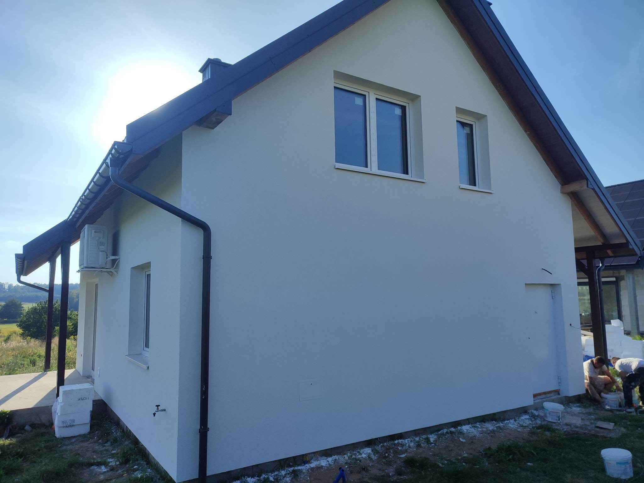 Świeżo otynkowana elewacja domu jednorodzinnego w jasnym odcieniu, widoczne okna z białymi ramami, fragment dachu z ciemną blachodachówką, pracownicy w trakcie wykończenia dolnej części ściany.