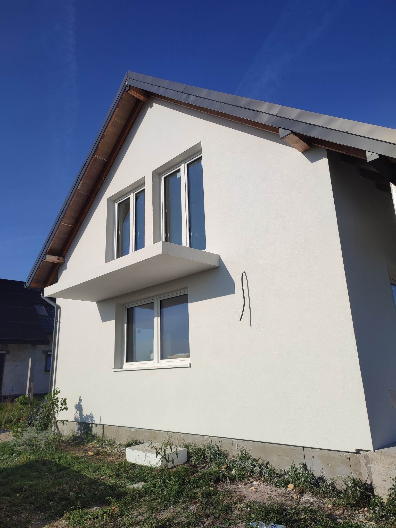 Nowoczesny dom jednorodzinny z jasną elewacją, widoczne okna z białymi ramami i wystający balkon, kabel zwisający ze ściany, błękitne niebo w tle.