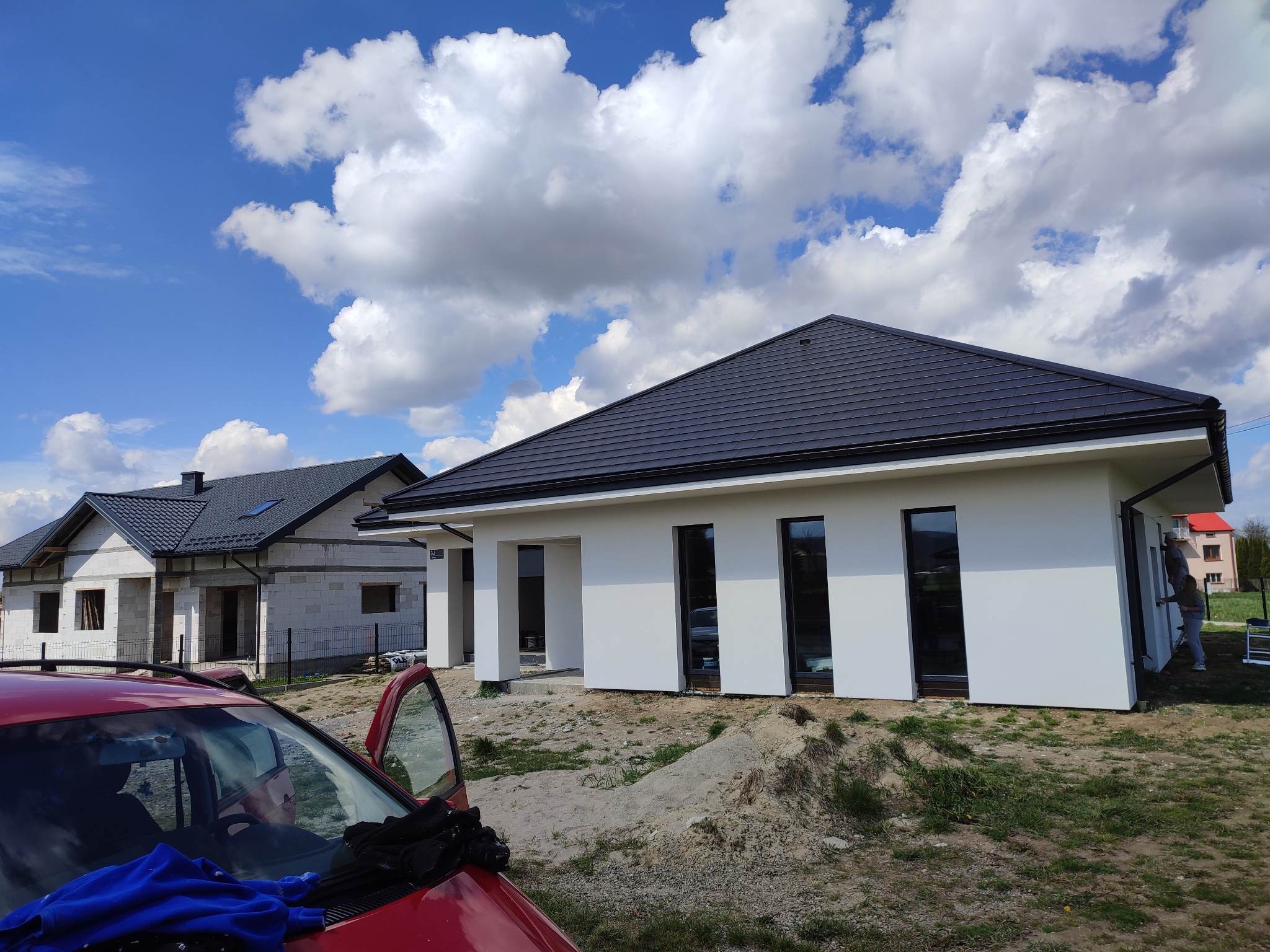 Nowoczesny, biały dom jednorodzinny z ciemną dachówką i wysokimi oknami, częściowo ukończony, z widocznym drugim budynkiem w stanie surowym i czerwonym samochodem z otwartymi drzwiami na pierwszym...