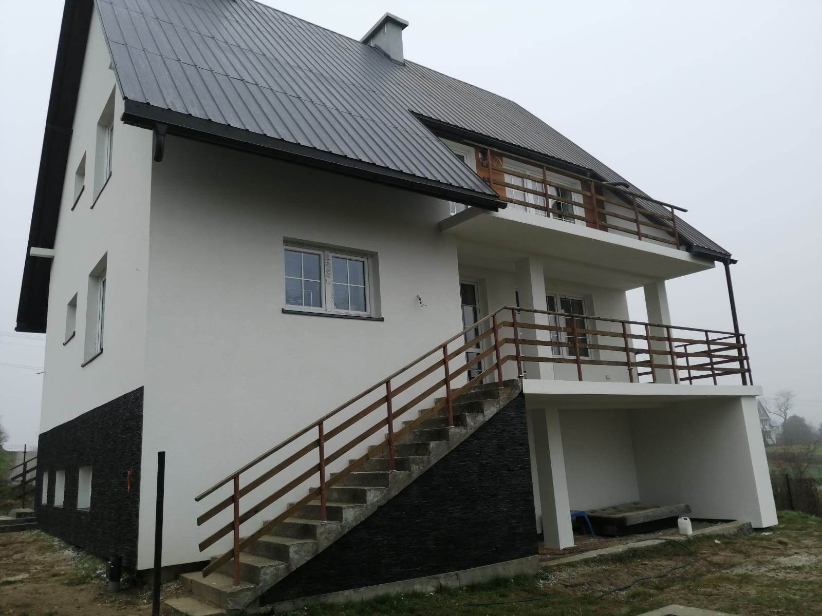 Nowoczesny dom z białą elewacją, fragmentem ściany wykończonym czarnym kamieniem dekoracyjnym, metalowym dachem i drewnianą balustradą balkonu.