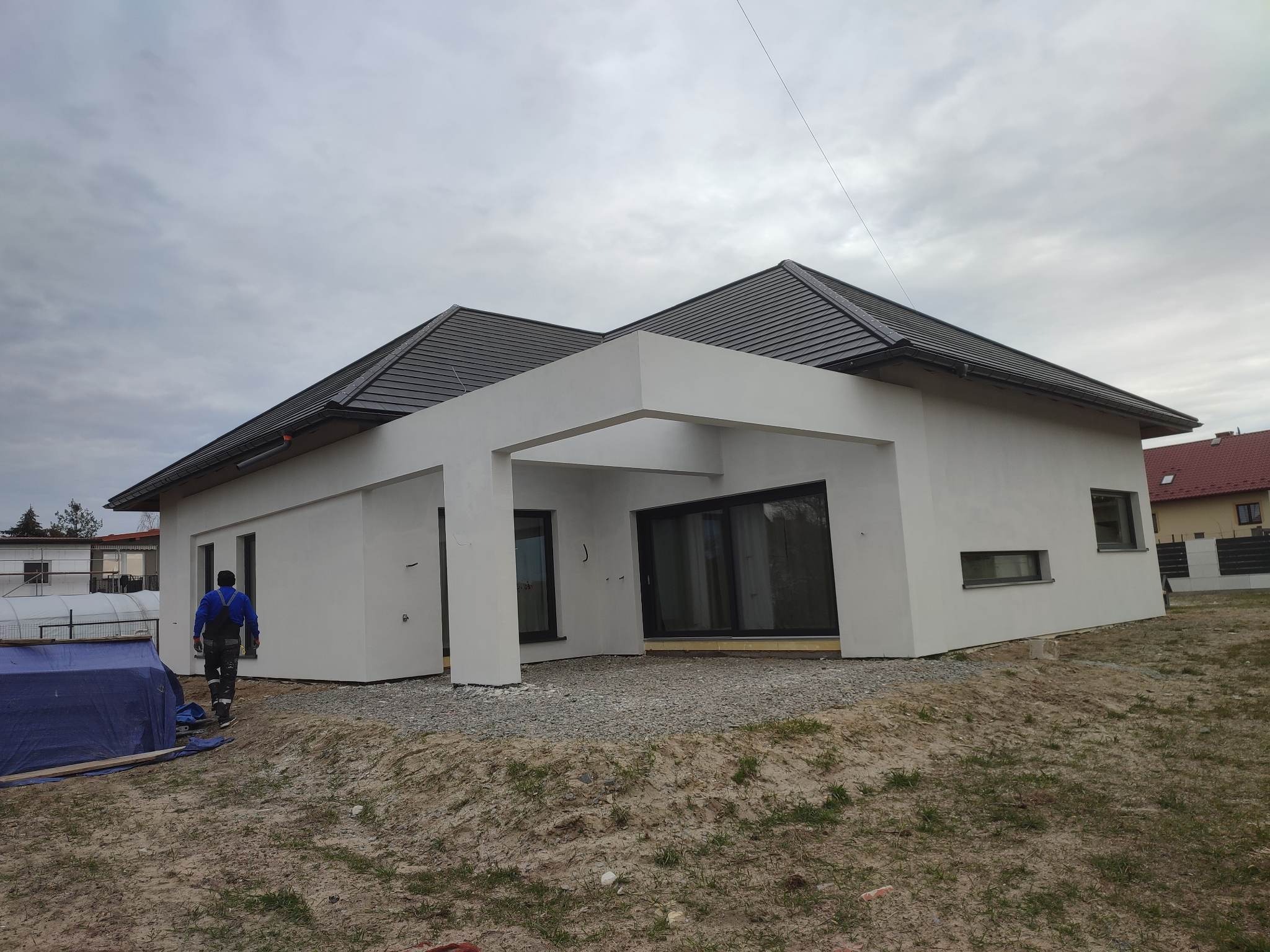 Nowoczesny dom jednorodzinny z białą elewacją i grafitowym dachem, widok z zewnątrz, teren wokół w trakcie zagospodarowywania, pracownik budowlany w oddali.