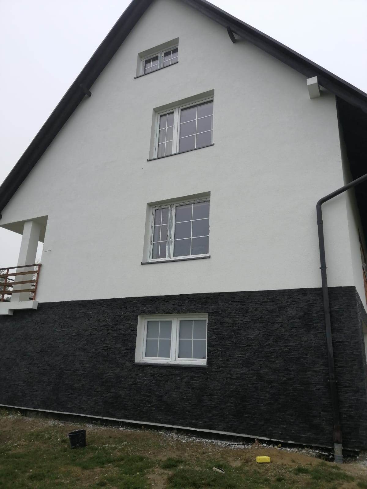 Dwukolorowa elewacja domu z białym tynkiem powyżej i ciemnoszarą okładziną imitującą kamień poniżej, widoczne okna z białymi ramami i balkon z drewnianą balustradą.