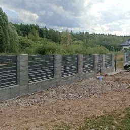 Nowoczesne ogrodzenie z poziomych, ciemnych paneli zamontowane na betonowych słupkach i podmurówce, widok z zewnątrz posesji, w tle dom i zieleń.