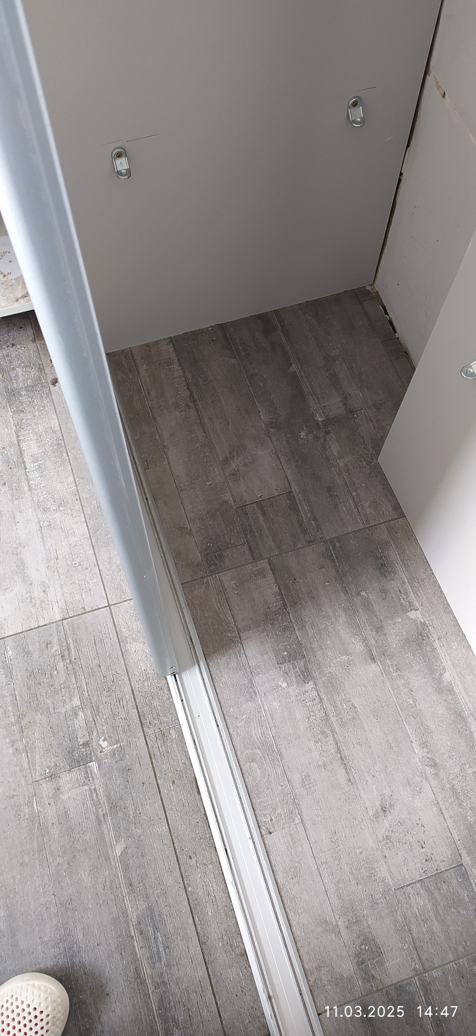 Szara, drewnopodobna podłoga w pokoju z widocznymi śladami po remoncie, fragmentem białej listwy przypodłogowej i fragmentem buta.