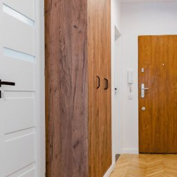 MW53 Sp. z o.o. - Korytarz z szafą w zabudowie o drewnianym dekorze, dopasowaną do podłogi z parkietu ułożonego w jodełkę. Białe drzwi po lewej stronie i brązowe drzwi wejściowe widoczne w tle.