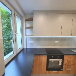 Niewielka kuchnia z drewnianymi szafkami dolnymi i jasnymi górnymi, czarnym blatem, płytą indukcyjną i piekarnikiem, widok na zieleń przez okno.