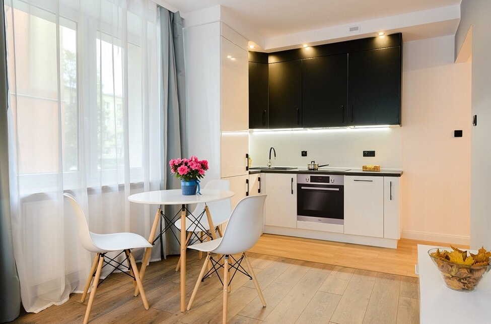 Mała, jasna kuchnia z czarnymi szafkami górnymi i białymi dolnymi, okrągły stół z bukietem różowych kwiatów, trzy białe krzesła z drewnianymi nogami, parkiet w odcieniach beżu i szarości.