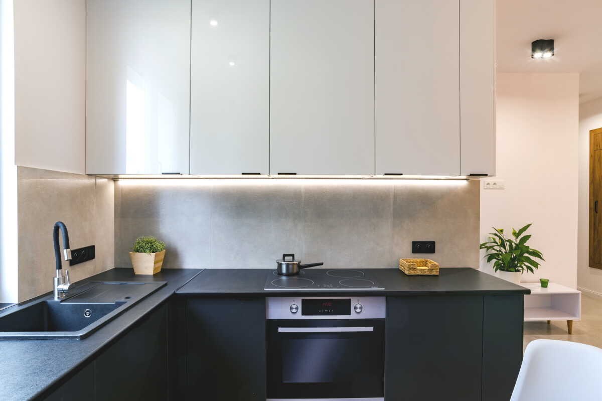 Nowoczesna kuchnia w odcieniach szarości i bieli z czarnym blatem, oświetleniem podszafkowym LED, zlewozmywakiem z czarną baterią i płytą indukcyjną z piekarnikiem. Na blacie stoi mały garnek, a...