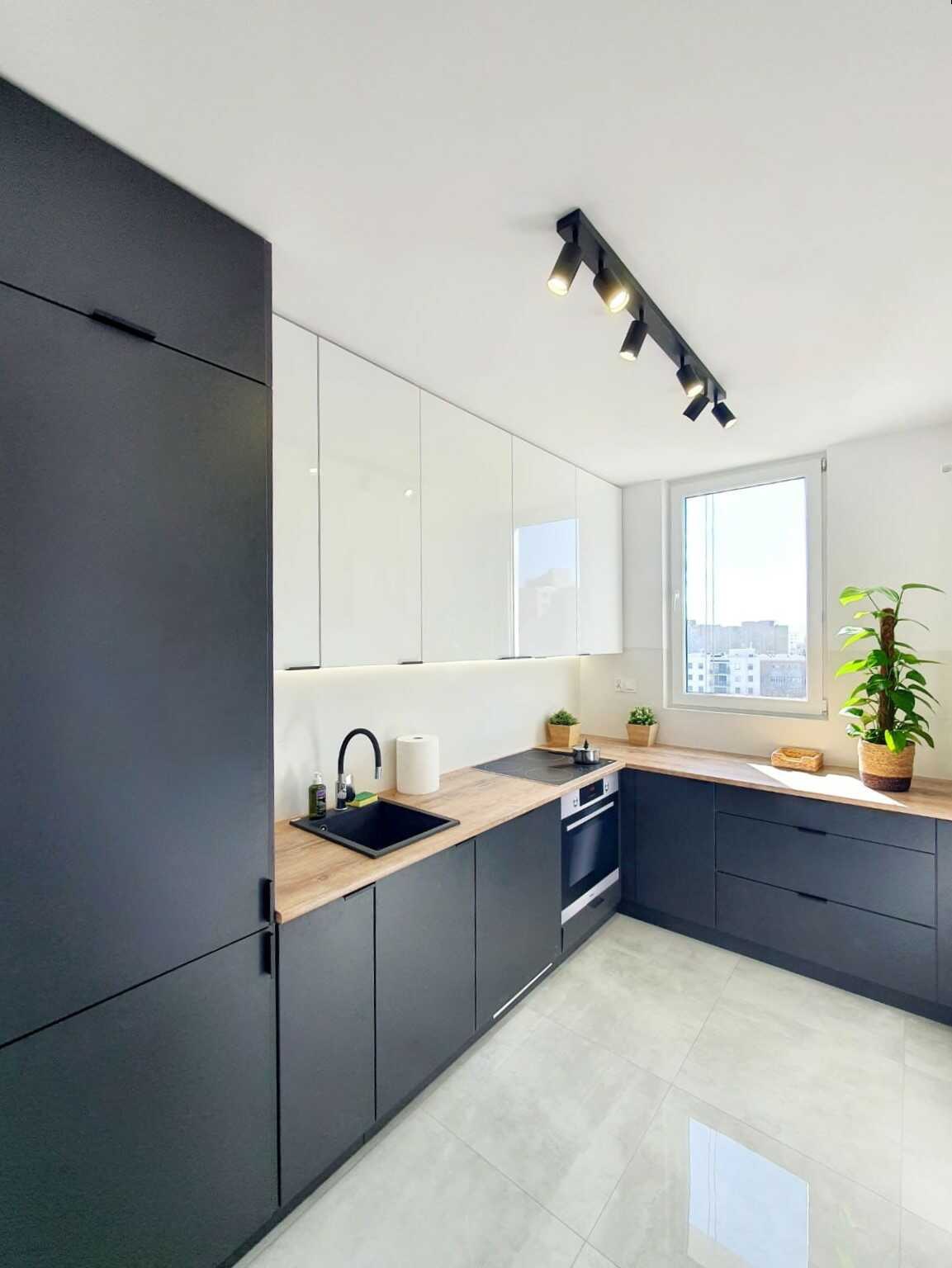 Dwukolorowa kuchnia z ciemnoszatynowymi dolnymi szafkami i jasnymi górnymi, drewniany blat, czarny zlew i bateria, oświetlenie punktowe na suficie, widok na miasto przez okno, roślina doniczkowa...