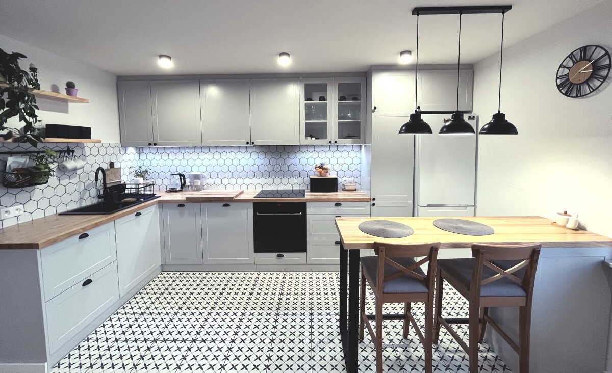 Jasna kuchnia z szarymi frontami, drewnianymi blatami, czarną armaturą i wzorzystą podłogą. Nad wyspą kuchenną wiszą trzy czarne lampy. Na ścianie wisi zegar.
