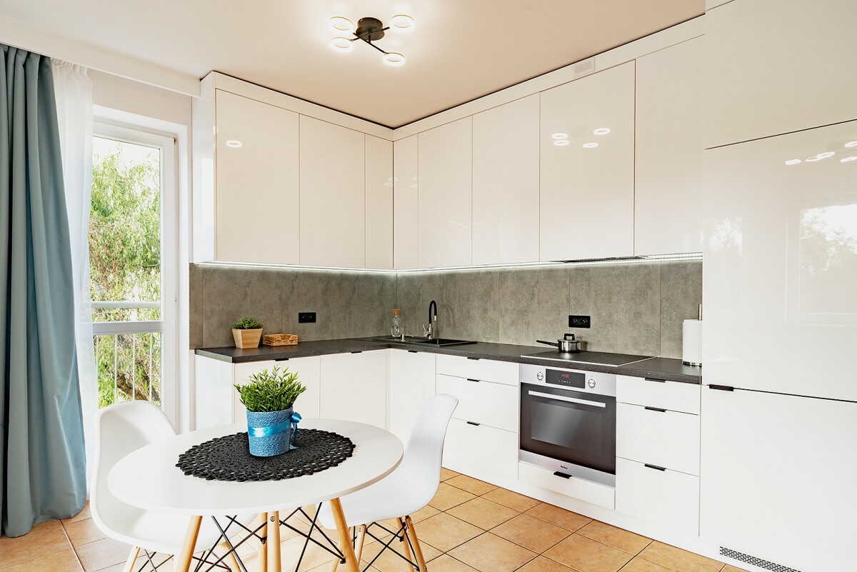 Nowoczesna kuchnia w kształcie litery L z białymi, lakierowanymi frontami na wysoki połysk, szarym blatem i podświetleniem LED pod górnymi szafkami, uzupełniona okrągłym stołem z rośliną...