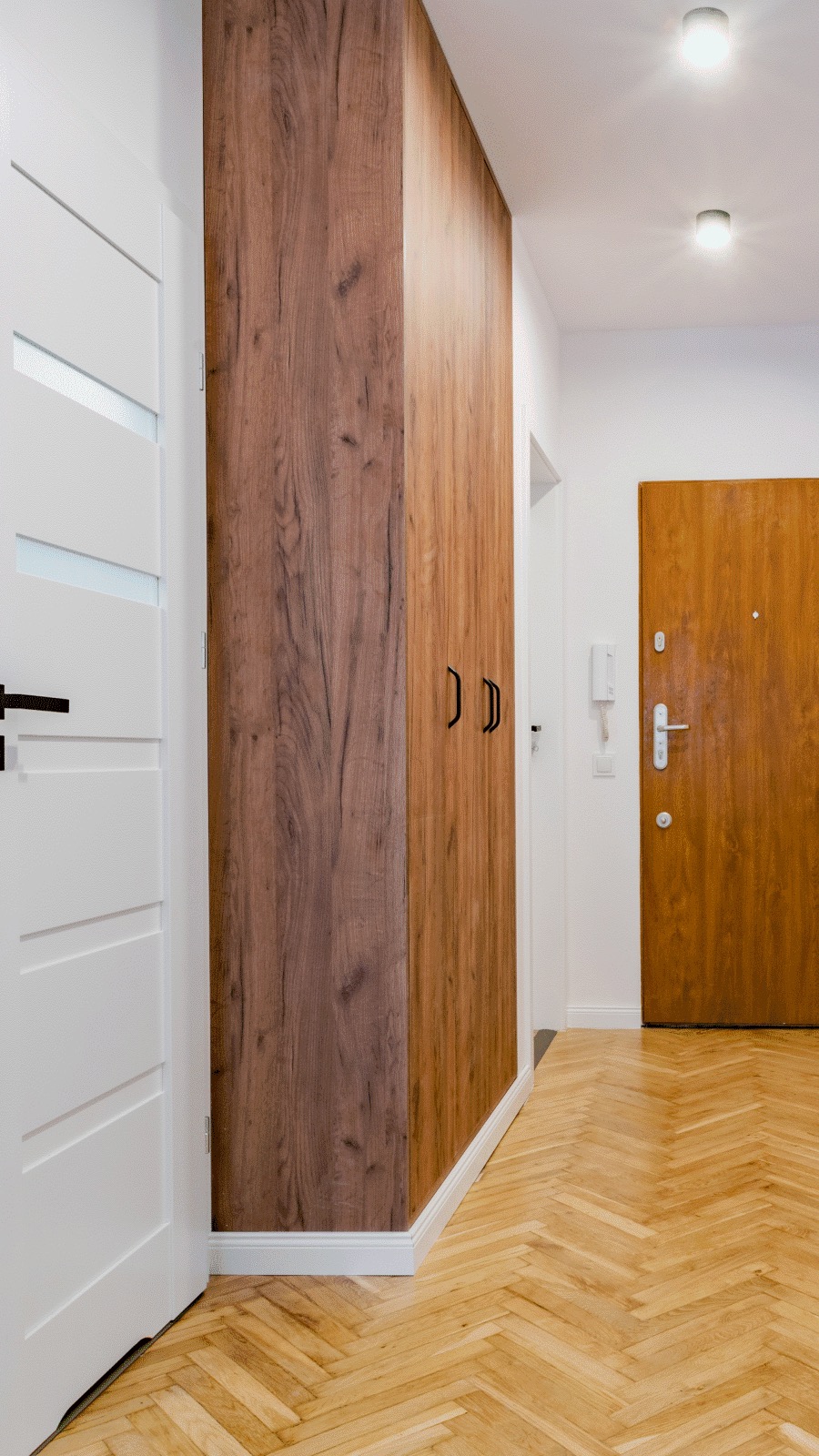 Korytarz z szafą w zabudowie o drewnianym dekorze, dopasowaną do podłogi z parkietu ułożonego w jodełkę. Białe drzwi po lewej stronie i brązowe drzwi wejściowe widoczne w tle.