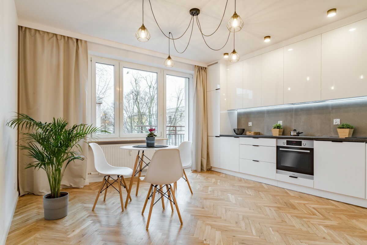 Jasna kuchnia z białymi szafkami na wysoki połysk, czarnym blatem, parkietem w jodełkę i okrągłym stołem z białymi krzesłami. Nad stołem zwisają dekoracyjne lampy. W oknie widok na drzewa.