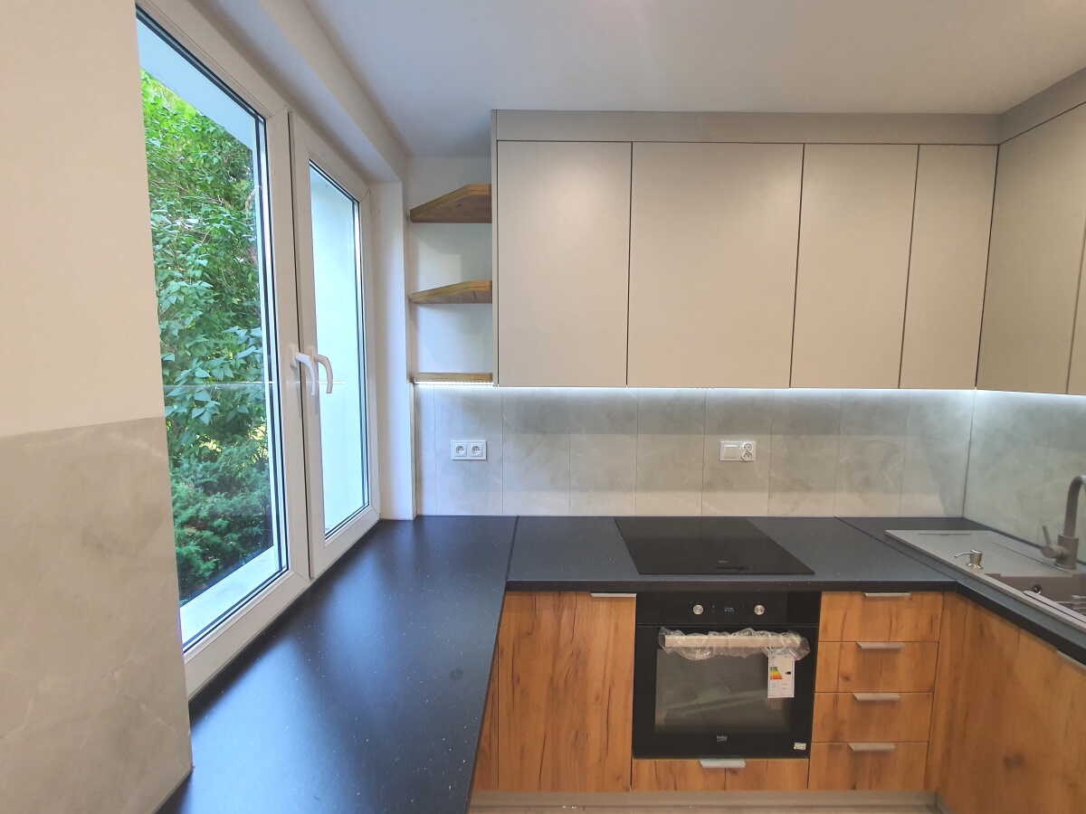 Niewielka kuchnia z drewnianymi szafkami dolnymi i jasnymi górnymi, czarnym blatem, płytą indukcyjną i piekarnikiem, widok na zieleń przez okno.