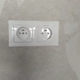 Podwójne, białe gniazdko elektryczne typu francuskiego zamontowane na ścianie z widocznymi śladami niedawnego malowania lub tapetowania.