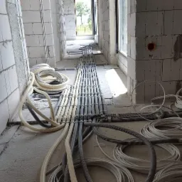 Instalacja elektryczna w surowym stanie: korytarz z ułożonymi rzędami szarych i białych peszli na podłodze, ściany z betonu komórkowego, widoczne puszki instalacyjne.