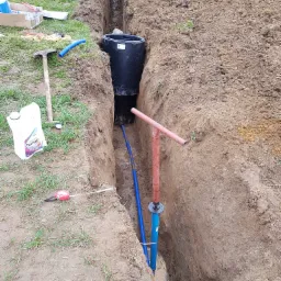 Głęboki wykop w ziemi z widoczną czarną studzienką kanalizacyjną, niebieskimi rurami i czerwoną rurą pionową z uchwytem T, obok leżą narzędzia i kanister z płynem.