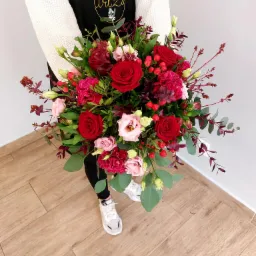 Osoba w czarnym fartuchu z logo 'Kawa i Kwiaty Art Zu' trzyma okazały bukiet czerwonych i różowych róż, goździków oraz eukaliptusa na tle drewnianej podłogi i białej ściany.