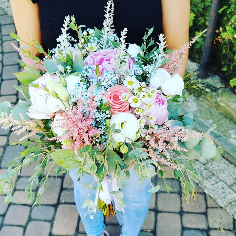 Osoba trzyma w rękach duży, bogaty bukiet z różowych i białych piwonii, róż, błękitnych gipsówek, białych eustom i zielonych eukaliptusów, przewiązany delikatną koronkową wstążką.