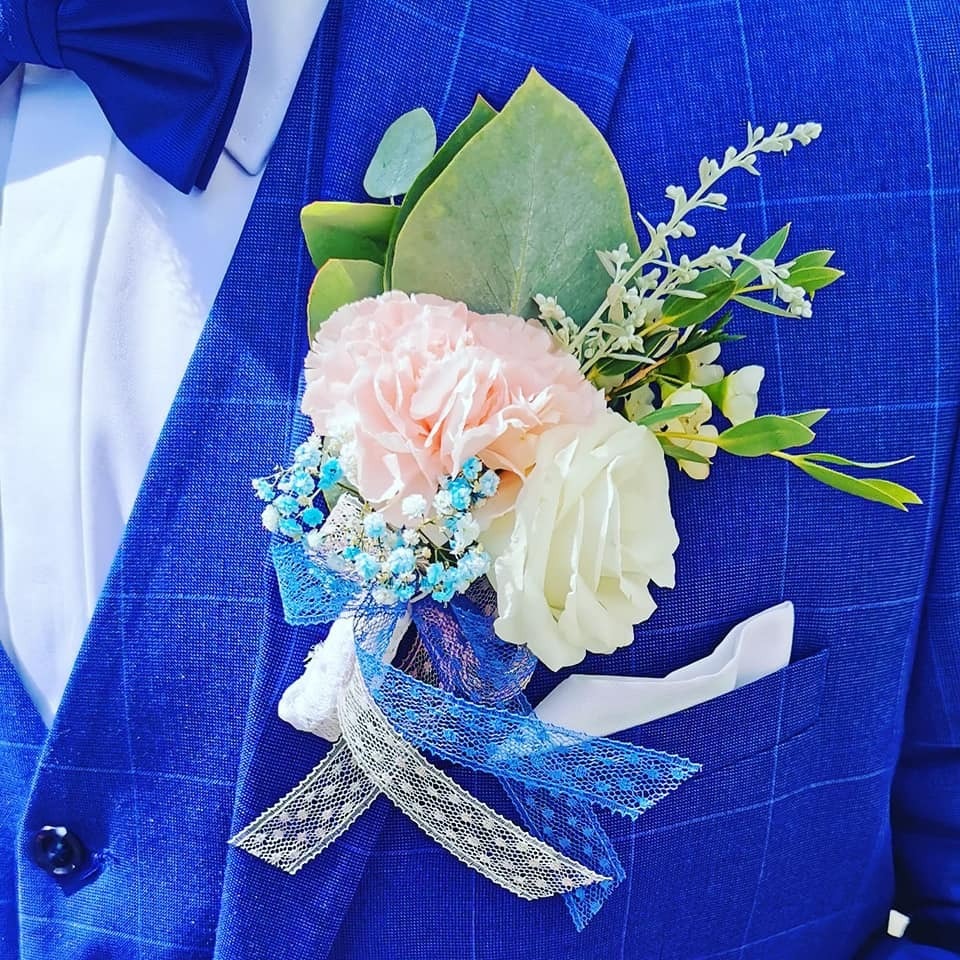 Błękitny garnitur z butonierką z różowej i białej róży, gipsówki i eukaliptusa, ozdobioną koronkową wstążką i włożoną w kieszeń marynarki białą poszetką, uzupełniony błękitną muchą.