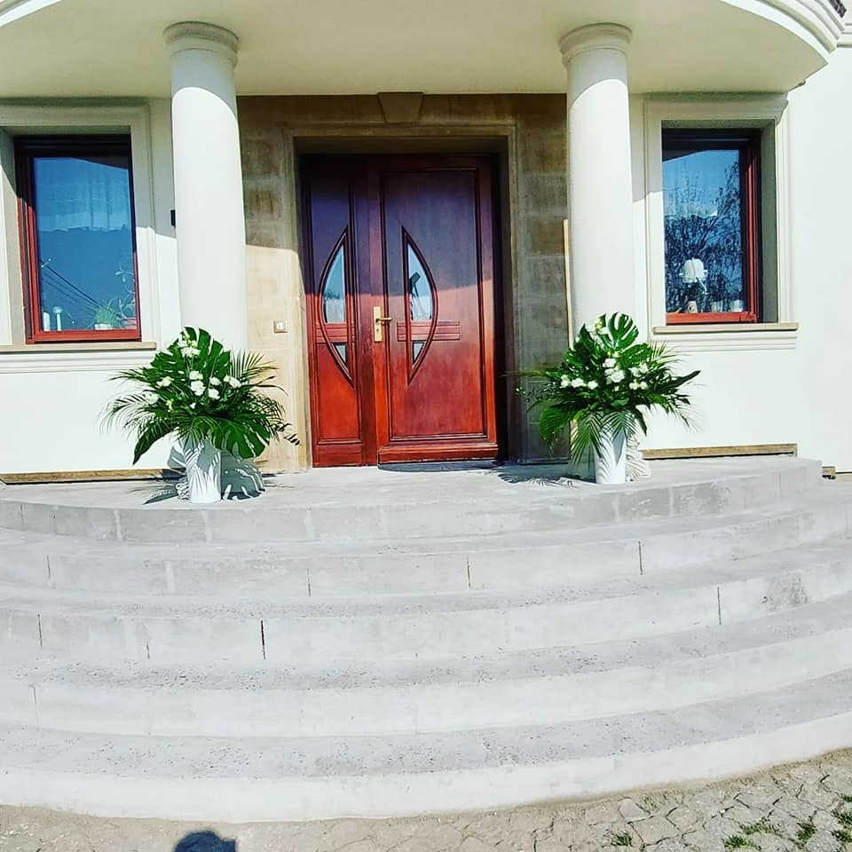 Dekoracja wejścia do domu z kwiatów i zielonych liści w białych wazonach, ustawiona po obu stronach drewnianych drzwi wejściowych.