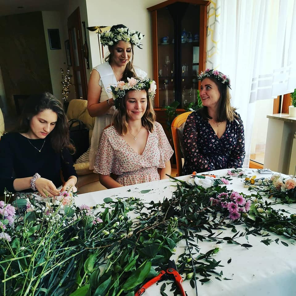 Kobiety w kwiecistych wiankach, jedna poprawia wianek innej, stół zastawiony kwiatami i zielenią, przygotowania do uroczystości.