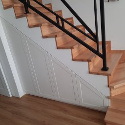 Rivej Sergii Yatsunov - Drewniane schody z czarną, metalową balustradą i zabudową szafek pod schodami. Wykończenie w drewnie i bieli, widok z dołu. Nowoczesny design i funkcjonalne rozwiązanie.