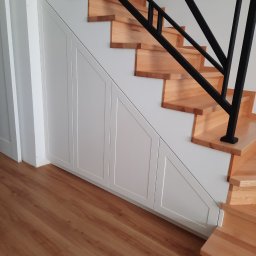 Rivej Sergii Yatsunov - Drewniane schody z czarną balustradą i zabudową pod schodami z białymi, frezowanymi drzwiczkami. Widok na fragment podłogi z paneli.