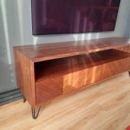 Rivej Sergii Yatsunov - Drewniana szafka RTV z fornirem ułożonym w jodełkę, osadzona na metalowych nóżkach, podwieszona pod telewizorem na szarej ścianie z listwami.