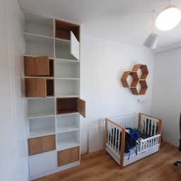 Rivej Sergii Yatsunov - Biała szafa wnękowa z drewnianymi akcentami w pokoju dziecięcym, obok łóżeczka. Widoczne półki i szafki z otwartymi drzwiczkami.