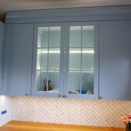 Rivej Sergii Yatsunov - Niebieska zabudowa kuchenna z przeszklonymi szafkami podświetlonymi LED, drewniany blat i białe płytki ułożone w jodełkę. Widoczne złote akcenty.