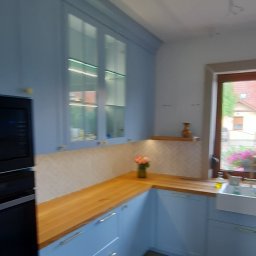 Rivej Sergii Yatsunov - Błękitna kuchnia z drewnianym blatem i przeszklonymi szafkami. Na blacie wazon z kwiatami, w tle okno z widokiem na zieleń. Nowoczesny design, jasne wnętrze.