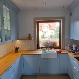 Rivej Sergii Yatsunov - Jasna kuchnia z błękitnymi szafkami, drewnianym blatem i białym zlewem farmerskim. W tle okno z widokiem na dom i kwiaty, płytki w jodełkę na ścianie.