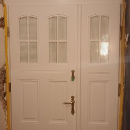 Rivej Sergii Yatsunov - Białe dwuskrzydłowe drzwi wewnętrzne z szybkami w kratkę i ozdobną, złotą klamką, zamontowane w ościeżnicy zabezpieczonej żółtą taśmą malarską.