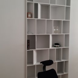 Rivej Sergii Yatsunov - Biała, wysoka szafa z licznymi półkami, na których znajdują się książki i dekoracje. U dołu widoczne krzesło biurowe. Minimalistyczny design.