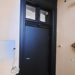 Rivej Sergii Yatsunov - Czarne, drewniane drzwi z ozdobnymi detalami i górnym oknem, wkomponowane w klasyczną ramę. Widoczny fragment ściany z instalacją elektryczną.