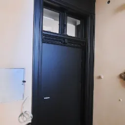 Czarne, drewniane drzwi z ozdobnymi detalami i górnym oknem, wkomponowane w klasyczną ramę. Widoczny fragment ściany z instalacją elektryczną.