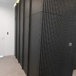 Nowoczesna, szara szafa wnękowa z lamelami i gładkimi frontami, wkomponowana w jasne wnętrze z widocznymi elementami instalacji elektrycznej.