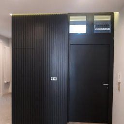 Rivej Sergii Yatsunov - Ciemna, gładka, drewniana ściana z pionowymi listwami i włącznikiem światła, połączona z ciemnymi drzwiami z nadświetlem w nowoczesnym wnętrzu.