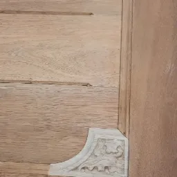 Detal wykończenia drewnianej szafy z dekoracyjnym, rzeźbionym narożnikiem. Widoczna struktura drewna i precyzja wykonania.