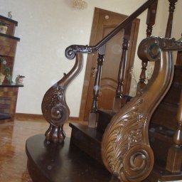Rivej Sergii Yatsunov - Eleganckie, ciemne schody drewniane z bogato rzeźbioną balustradą, widoczne detale zdobień i połysk lakieru. W tle drzwi i fragment ściany.