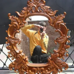 Ozdobne, drewniane lustro w bogato rzeźbionej ramie z motywami roślinnymi, odbijające postać fotografa z aparatem na tle ceglanej ściany i metalowej balustrady.