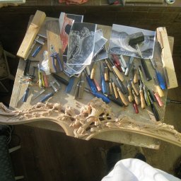 Rivej Sergii Yatsunov - Warsztat stolarski: Stół roboczy z narzędziami, dłutami, młotkiem i szablonami obok fragmentu ramy z dekoracyjnymi, rzeźbionymi elementami roślinnymi w jasnym drewnie.