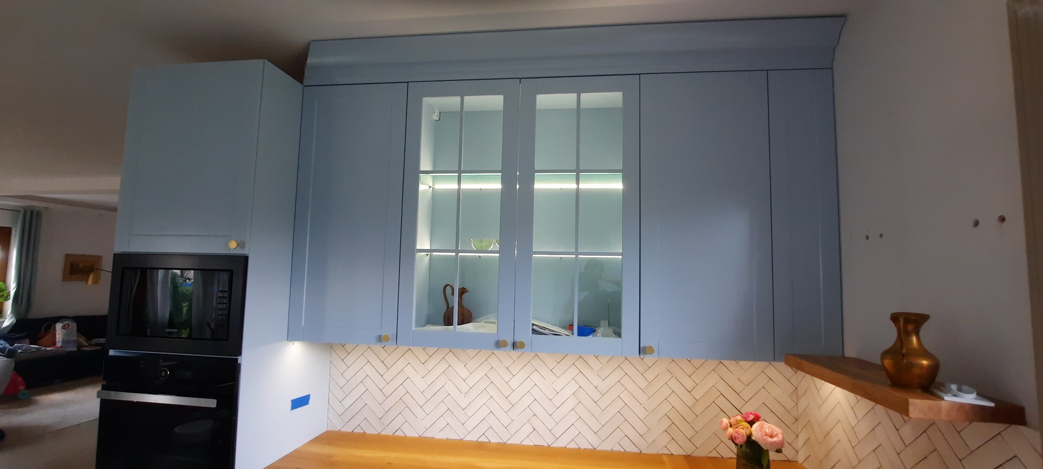 Niebieska zabudowa kuchenna z przeszklonymi szafkami podświetlonymi LED, drewniany blat i białe płytki ułożone w jodełkę. Widoczne złote akcenty.