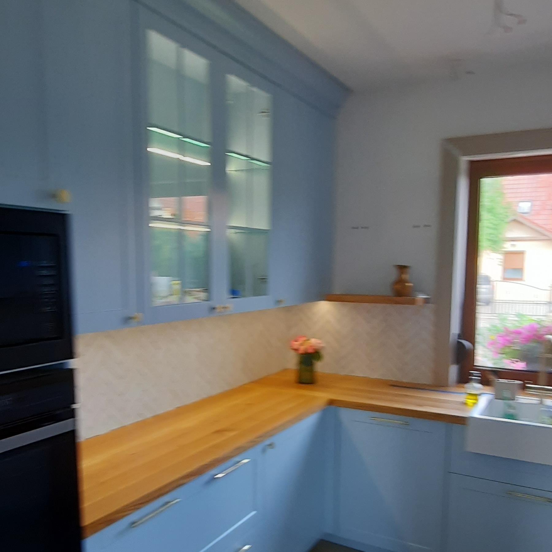 Błękitna kuchnia z drewnianym blatem i przeszklonymi szafkami. Na blacie wazon z kwiatami, w tle okno z widokiem na zieleń. Nowoczesny design, jasne wnętrze.