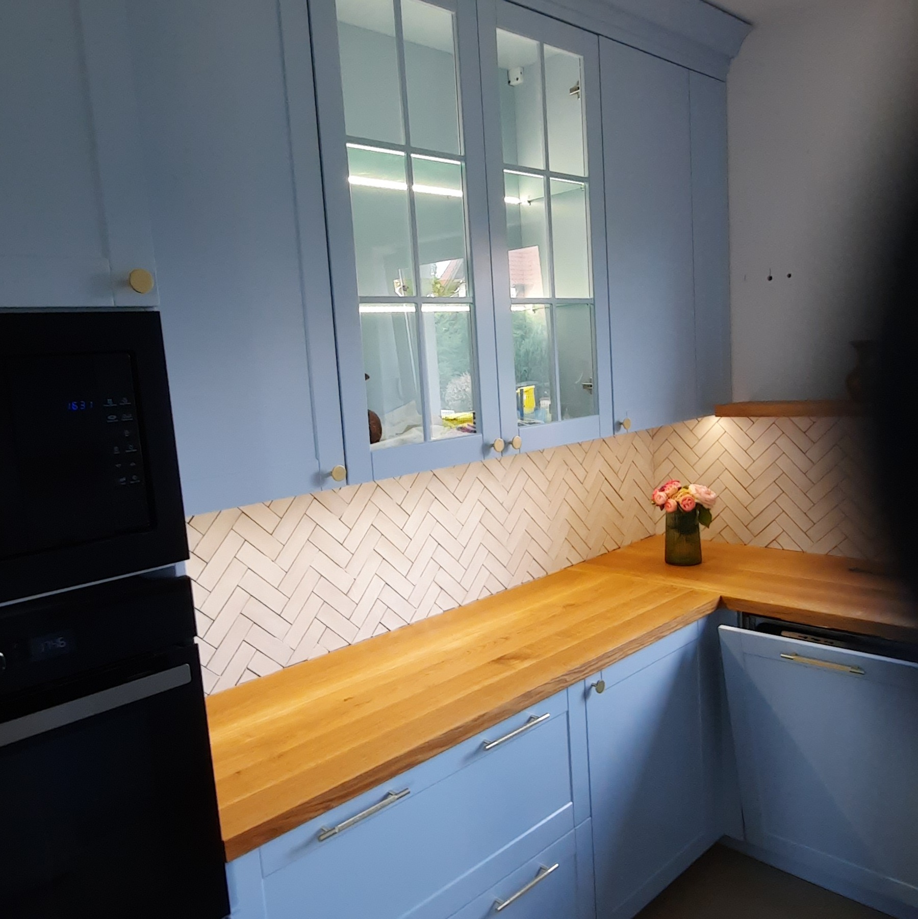 Błękitna kuchnia z drewnianym blatem, oświetleniem podszafkowym i szklanymi frontami górnych szafek, uzupełniona o dekoracyjny akcent w postaci wazonu z kwiatami.