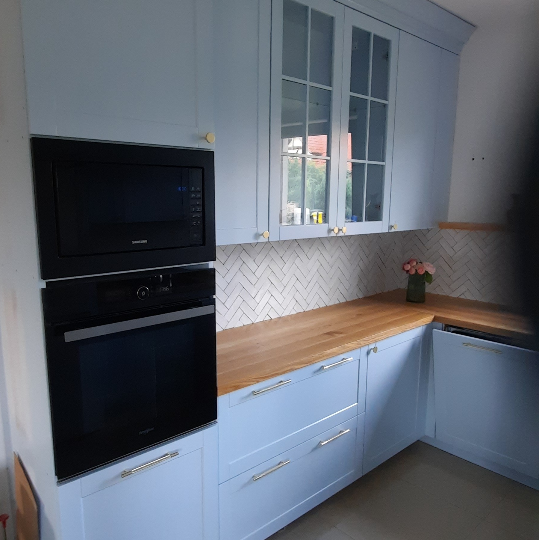 Jasna kuchnia z drewnianym blatem i białymi płytkami ułożonymi w jodełkę. W zabudowie piekarnik i mikrofalówka. Na blacie wazon z różowymi kwiatami.