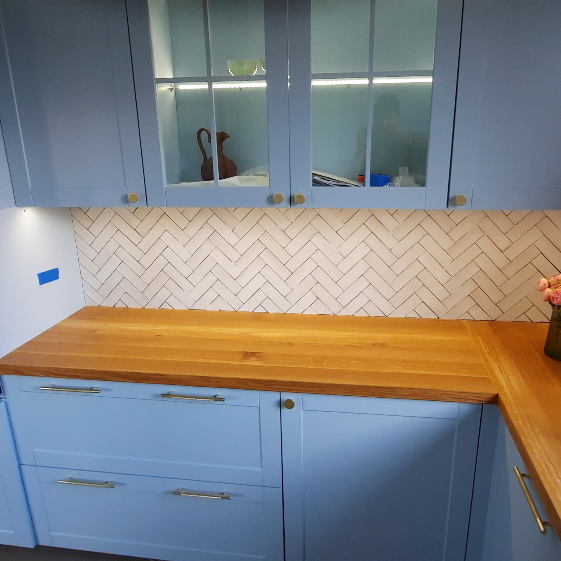 Jasna kuchnia: drewniany blat, błękitne szafki z złotymi uchwytami. Nad blatem białe płytki ułożone w jodełkę. Górne szafki przeszklone, podświetlone LED.